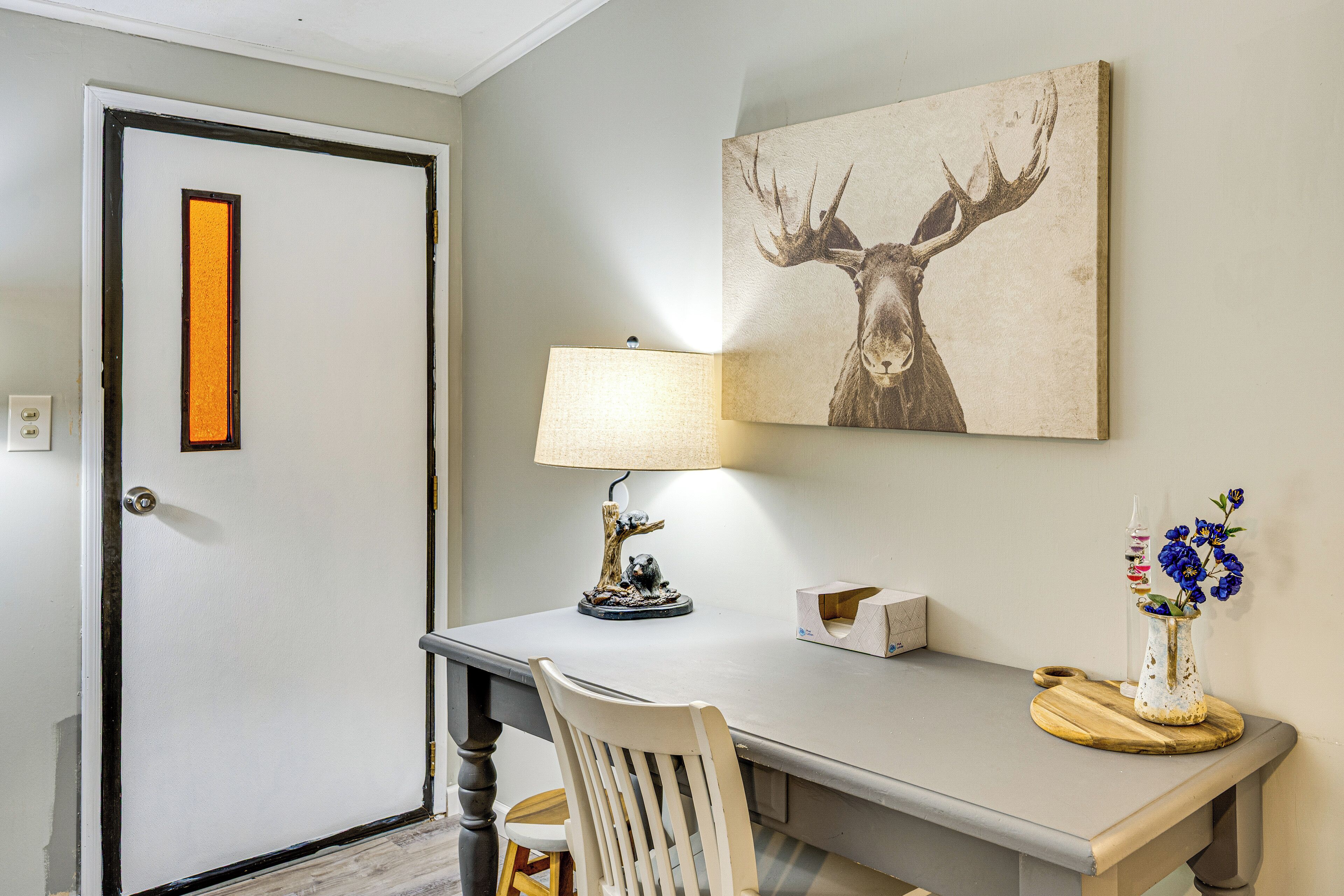 A room with a desk, a lamp, a chair, a vase with flowers, and a framed picture on the wall.
