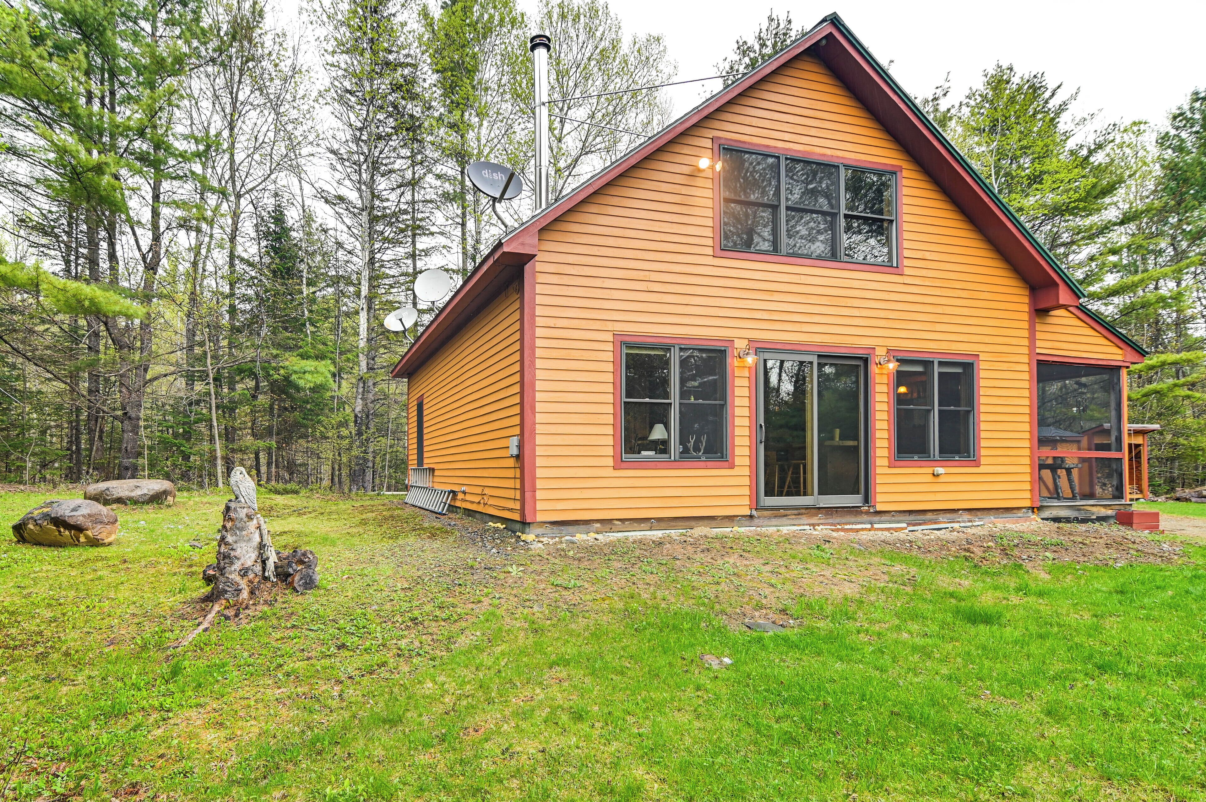 A two-story wooden cabin with large windows and a porch, surrounded by a forested area.