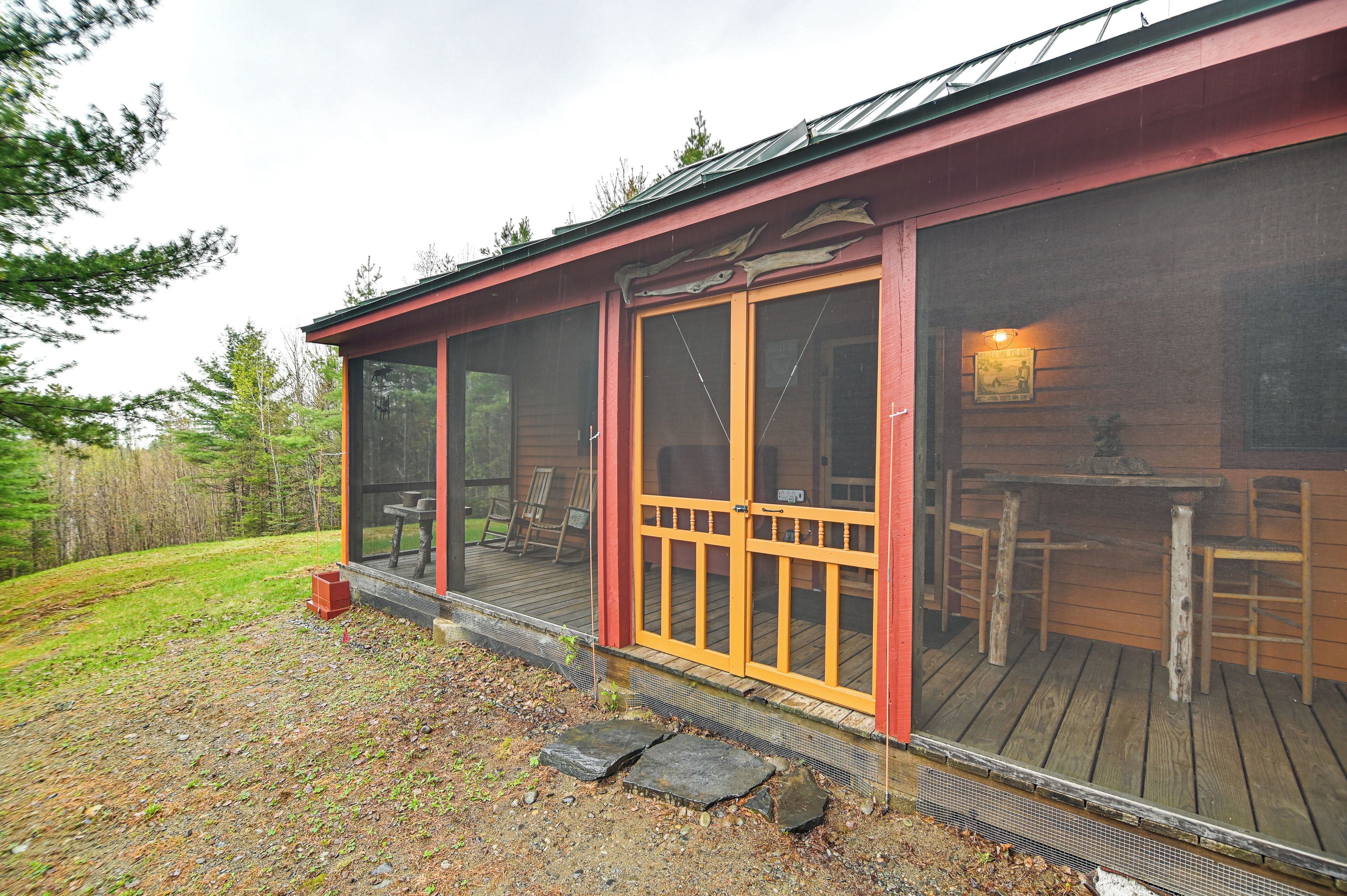 A wooden cabin with a covered porch, a dining area inside, and a view of the surrounding forest.