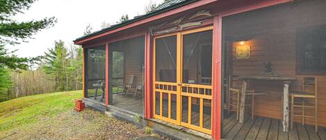 A wooden cabin with a covered porch, a dining area inside, and a view of the surrounding forest.
