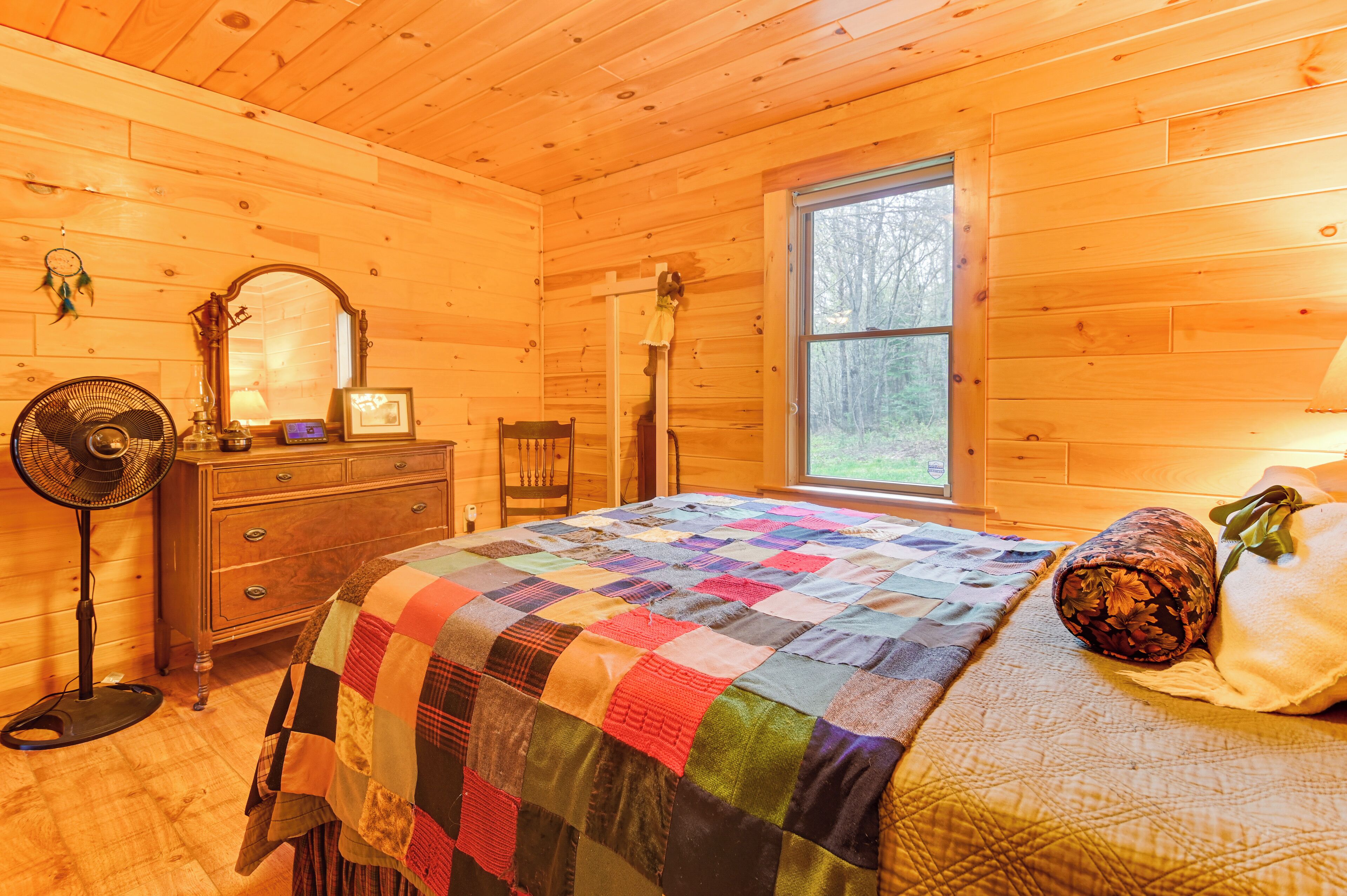 Ein gemütliches Schlafzimmer in einer Holzhütte, mit einer gesteppten Bettdecke, einem Schminktisch, einem Spiegel, einem Ventilator und einem Fenster mit Blick auf Bäume.
