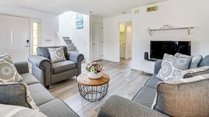 Una sala de estar moderna con sofás grises, una mesa de centro con un centro de flores, un televisor de pantalla plana y una escalera que lleva a otra habitación.