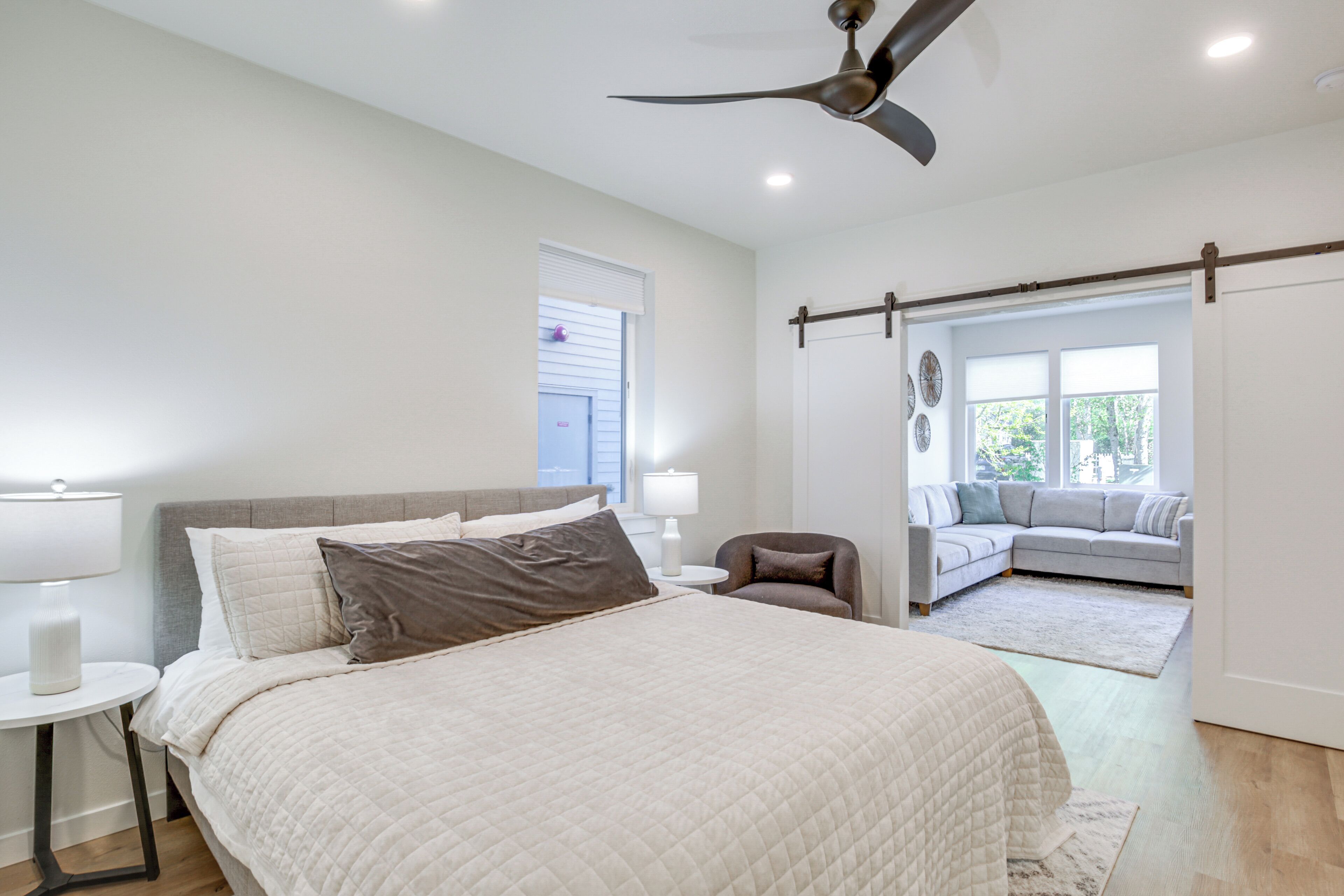 A bedroom with a bed, bedside tables, a ceiling fan, a sofa, and a view of the outdoors.