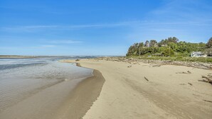 A beach with clear skies, a sandy shore, and a forested area with houses in the background. - Walk to Beaches: 'siletz Bay Bliss' Townhome (Lincoln City)