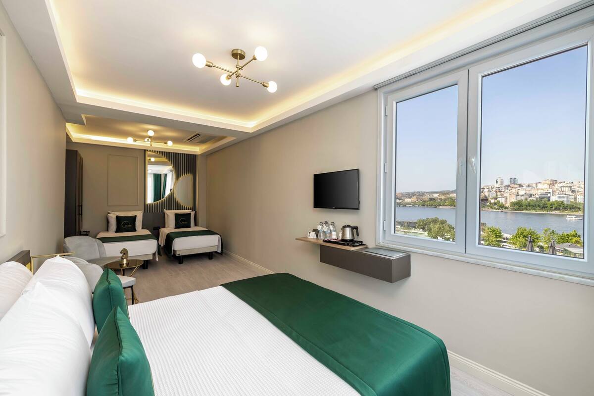 Une chambre d’hôtel moderne dotée d’un grand lit, d’un coin salon avec un canapé et des fauteuils, d’une télévision et offrant une vue sur le paysage urbain et l’eau.