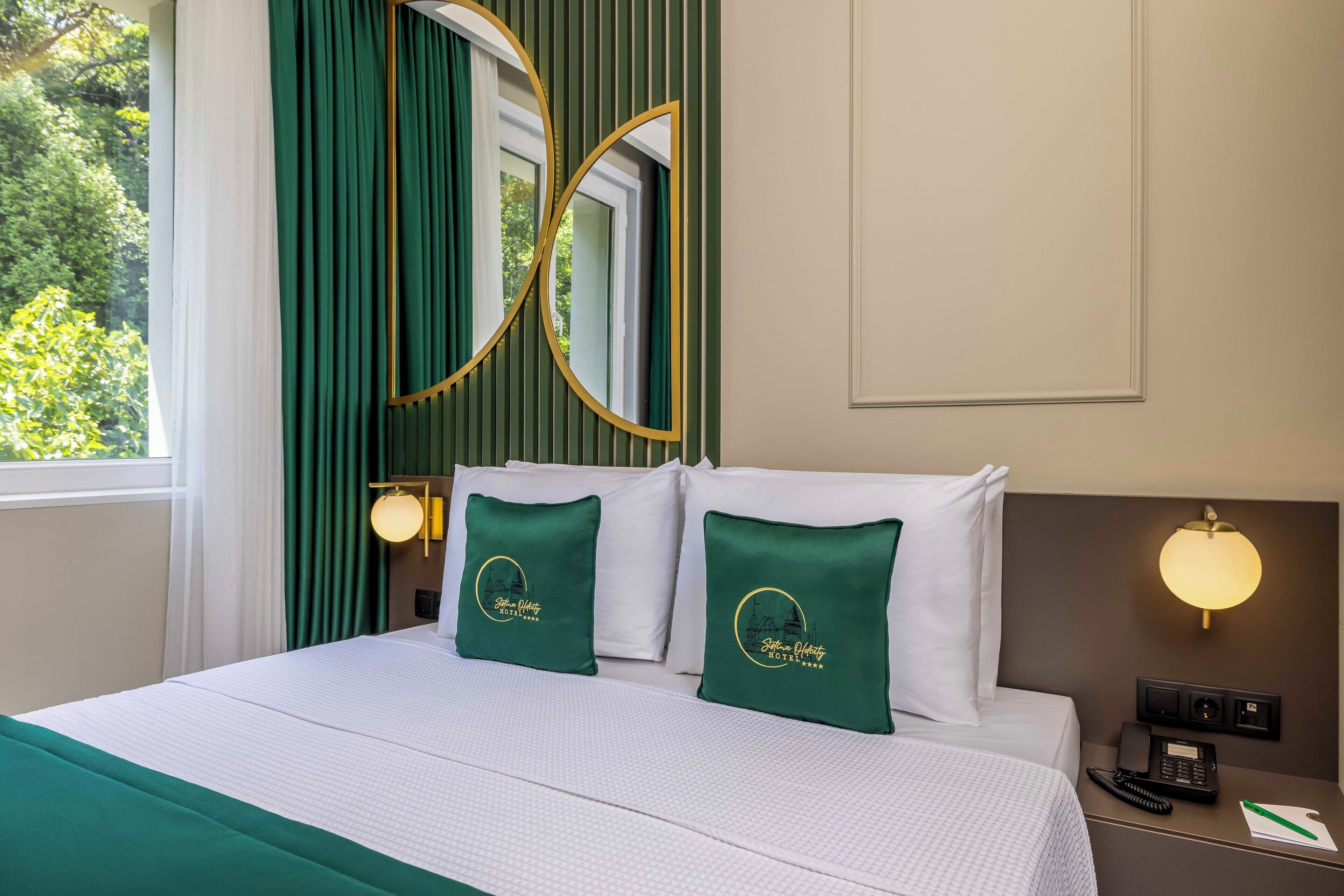 Habitación de hotel con cama, ropa de cama verde y blanca, mesita de noche con teléfono y lámpara, y ventana con cortinas.