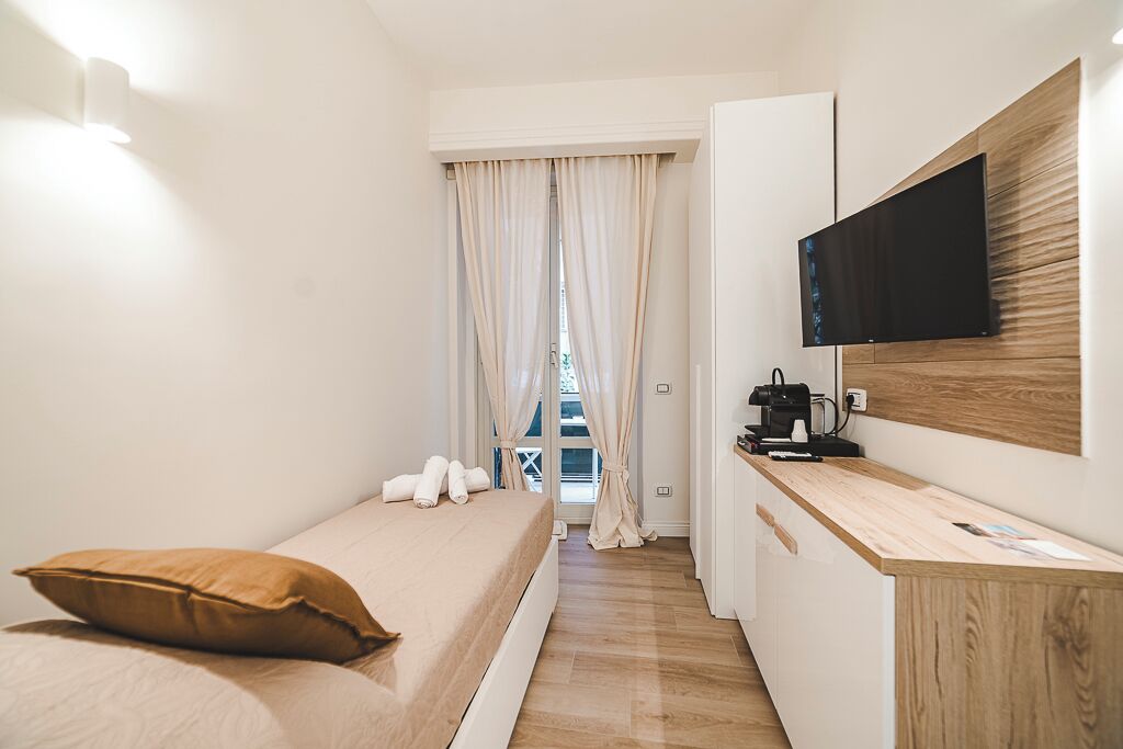 A compact hotel room with a single bed, a desk with a flat-screen TV, and a view through a window with curtains.