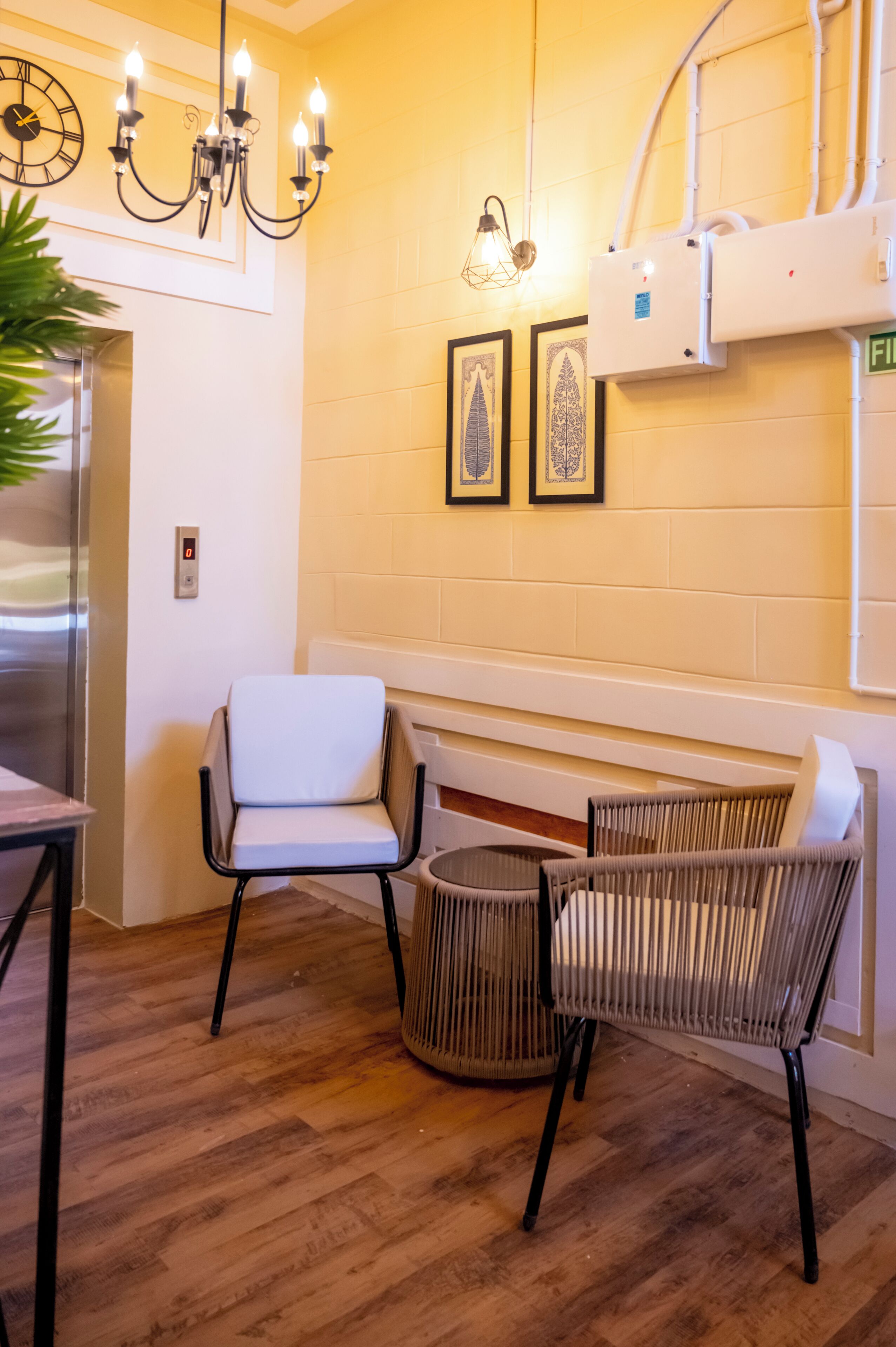 A room with a wooden floor, a white chair, a wicker chair, a wall-mounted light fixture, and a clock.