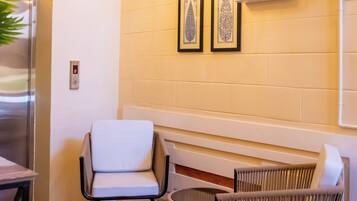 A room with a wooden floor, a white chair, a wicker chair, a wall-mounted light fixture, and a clock.