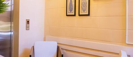 A room with a wooden floor, a white chair, a wicker chair, a wall-mounted light fixture, and a clock.
