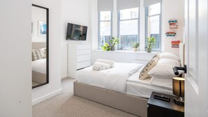 Ein modernes Schlafzimmer mit einem großen Bett, einem an der Wand montierten Fernseher, einem Fenster mit Pflanzen und einem Nachttisch mit Lampe.