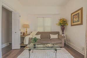 Un salon avec une table basse en verre, un canapé beige, un tapis blanc, une lampe et un vase rempli de fleurs.