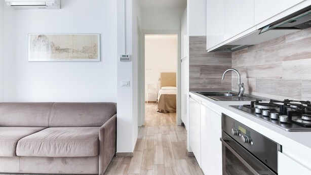 A modern kitchen with a gas stove, oven, and white cabinetry. A beige sofa is positioned in the living area, and a framed picture hangs on the wall. - Two-room Apartment Scola 12 (Gabicce Mare)