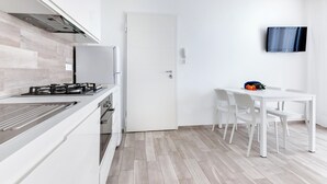 A modern kitchen with white cabinetry, a gas stove, and a dining area with white chairs and a table. - Two-room Apartment Scola 12 (Gabicce Mare)
