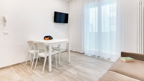 A modern living room with a white table, white chairs, a flat-screen TV mounted on the wall, a beige sofa, and a large window with sheer curtains. - Two-room Apartment Scola 12 (Gabicce Mare)