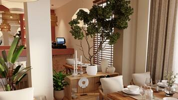Un coin repas avec une table en bois dressée pour un repas, entourée de chaises. On aperçoit une plante en pot sur la table, et une grande fenêtre avec des stores au fond.