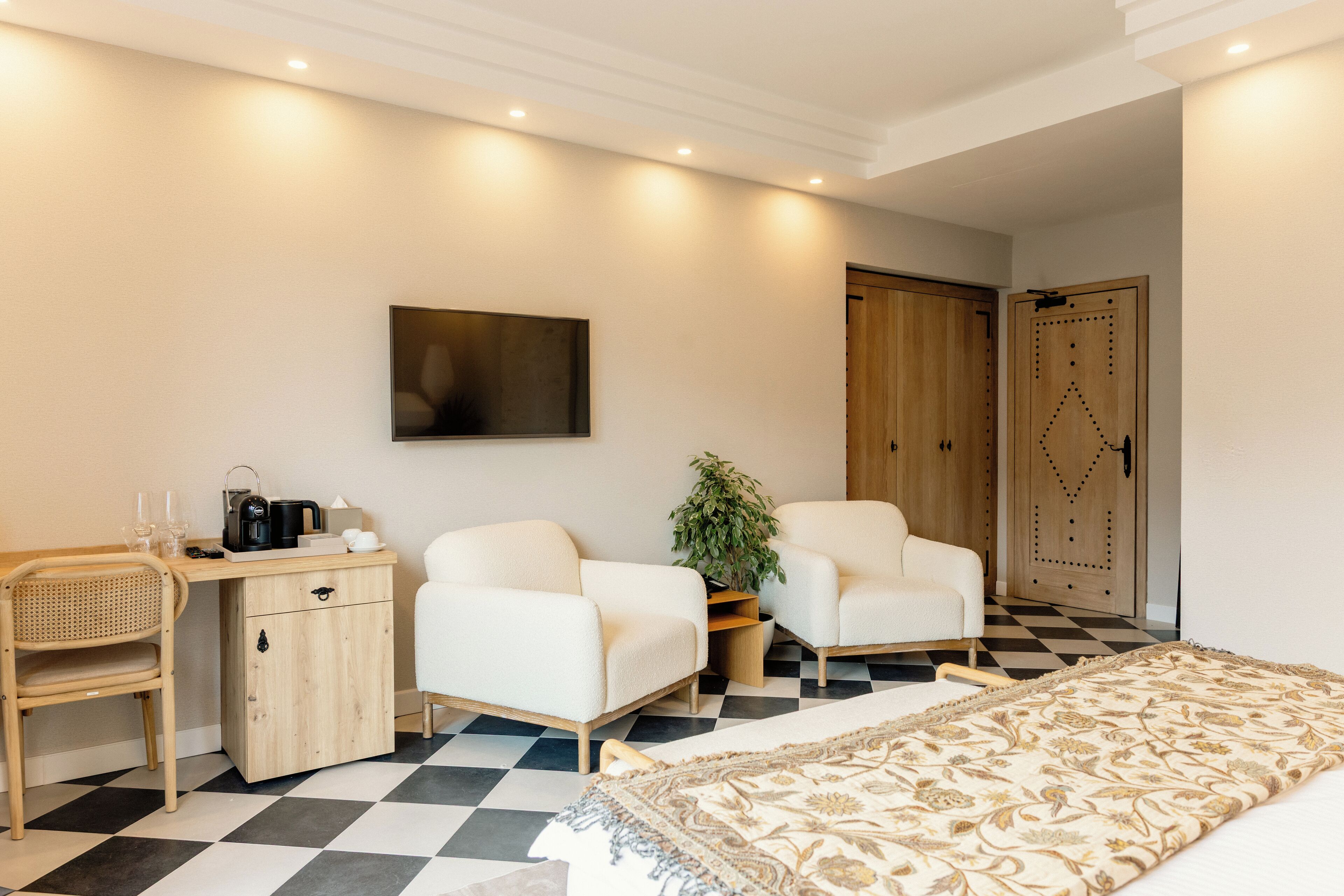 Habitación de hotel con suelo a cuadros, cama con cubrecama estampado, escritorio con cafetera, dos sillones blancos, planta en maceta y armario con puerta decorativa.