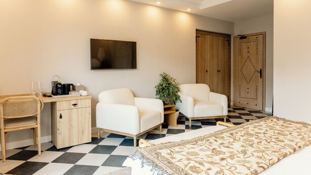 A hotel room with a checkered floor, a bed with a patterned bedspread, a desk with a coffee maker, two white armchairs, a potted plant, and a wardrobe with a decorative door. - Adiya Signature Hotel (Kotor)