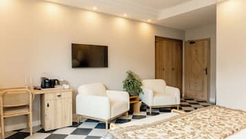 Habitación de hotel con suelo a cuadros, cama con cubrecama estampado, escritorio con cafetera, dos sillones blancos, planta en maceta y armario con puerta decorativa.
