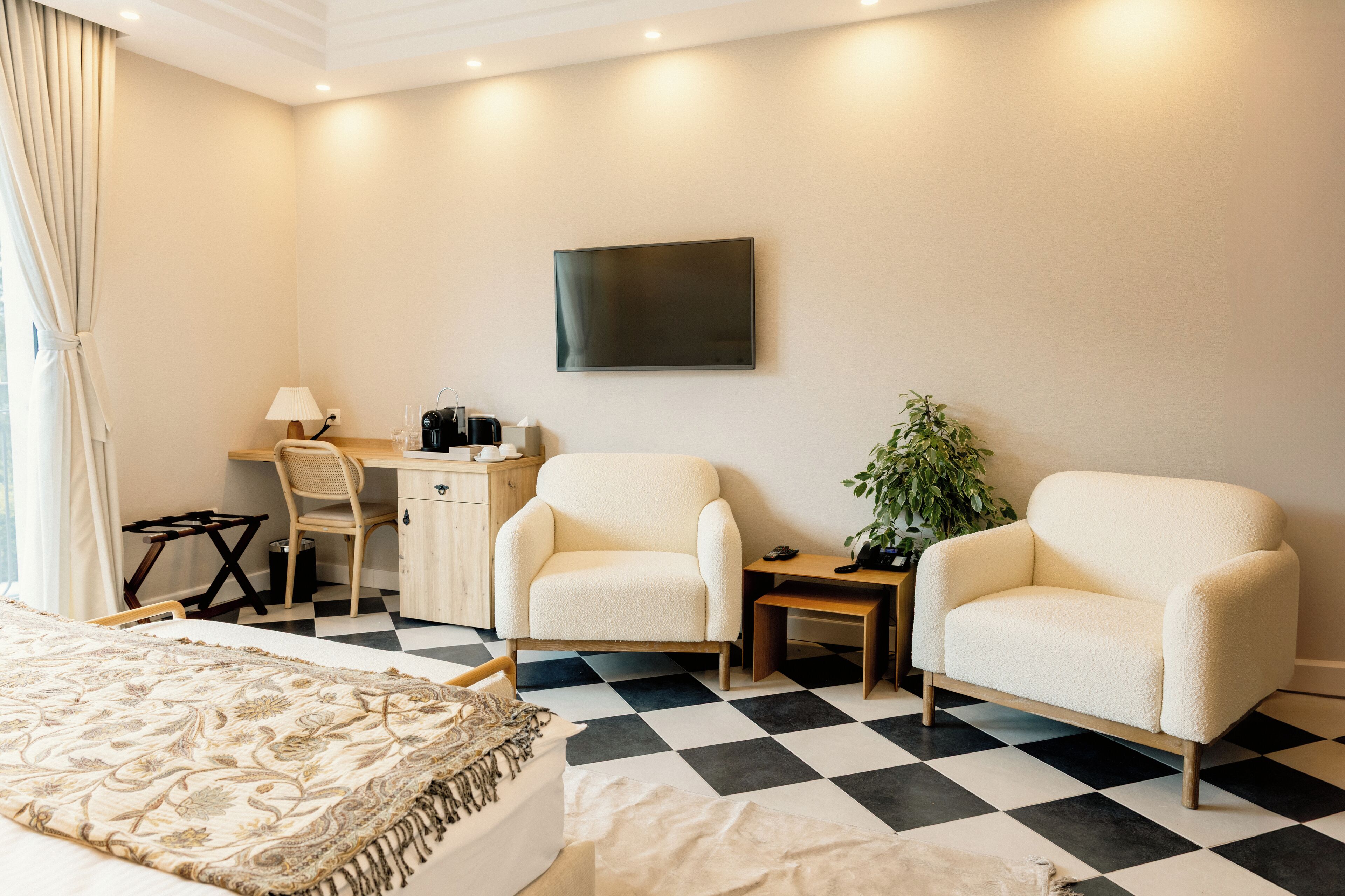 Een hotelkamer met een geruit vloerkleed, een bed met een bedspreide met dessin, twee fauteuils, een bureau met een stoel, een televisie en een potplant.