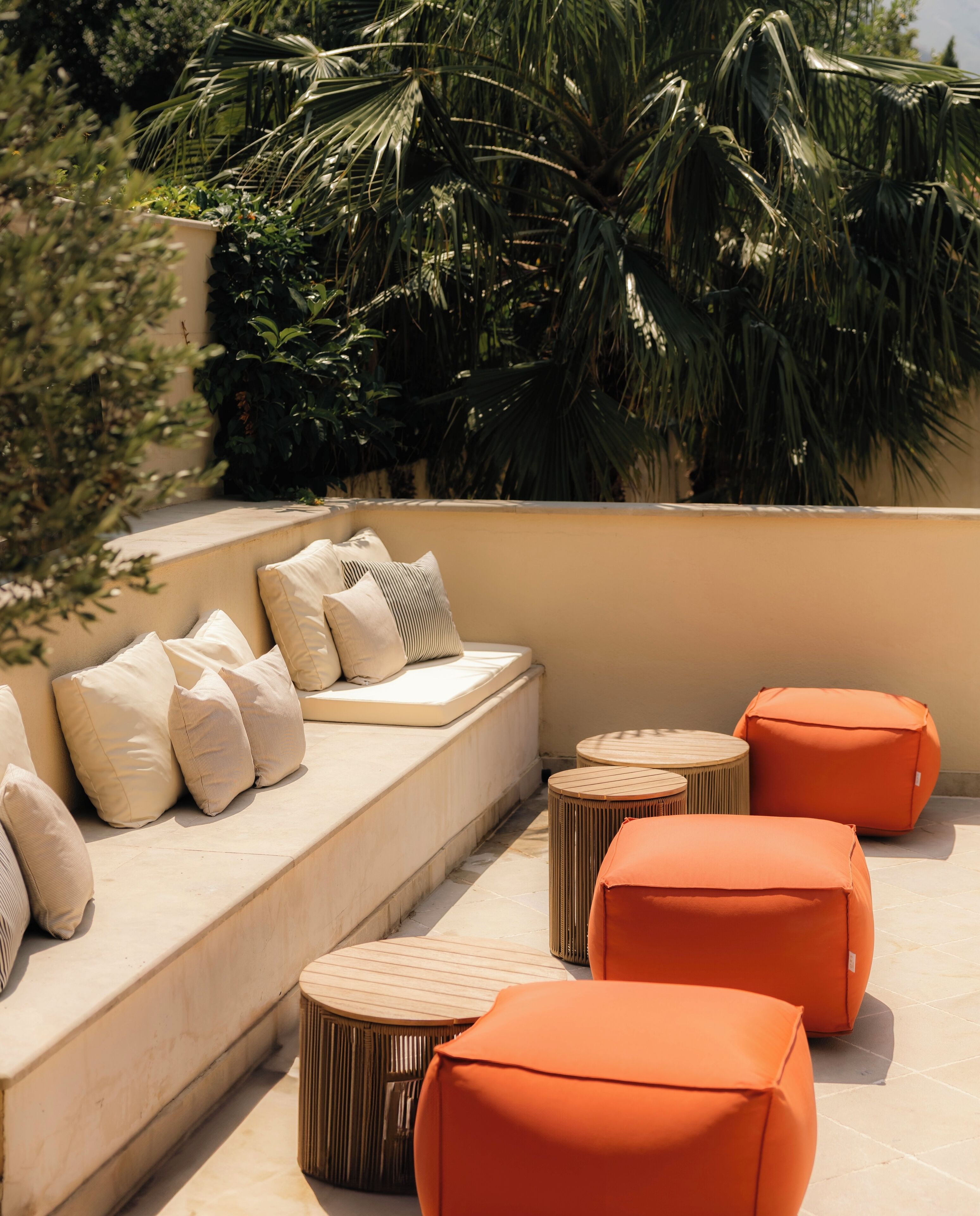 A sunlit outdoor seating area with a sofa and ottomans, surrounded by lush greenery.