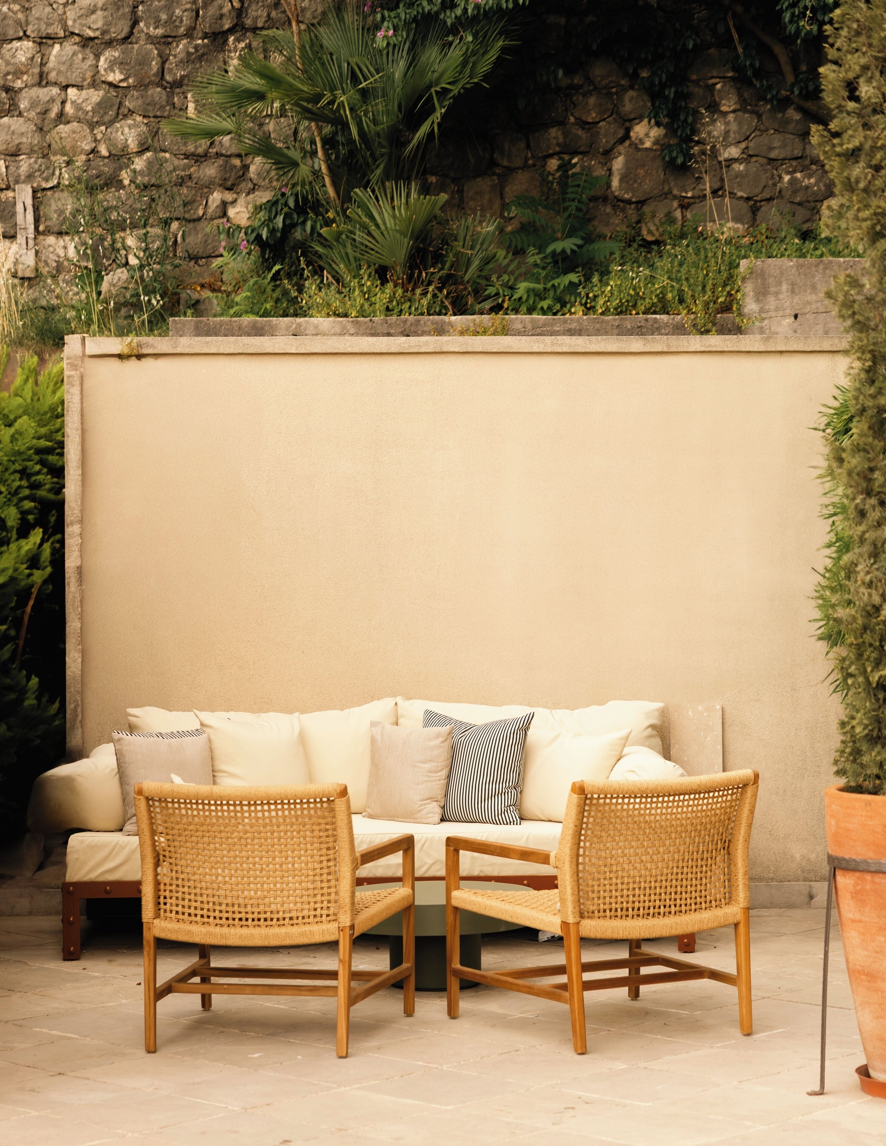 A beige sofa with two wicker chairs, a stone wall, and greenery in the background.