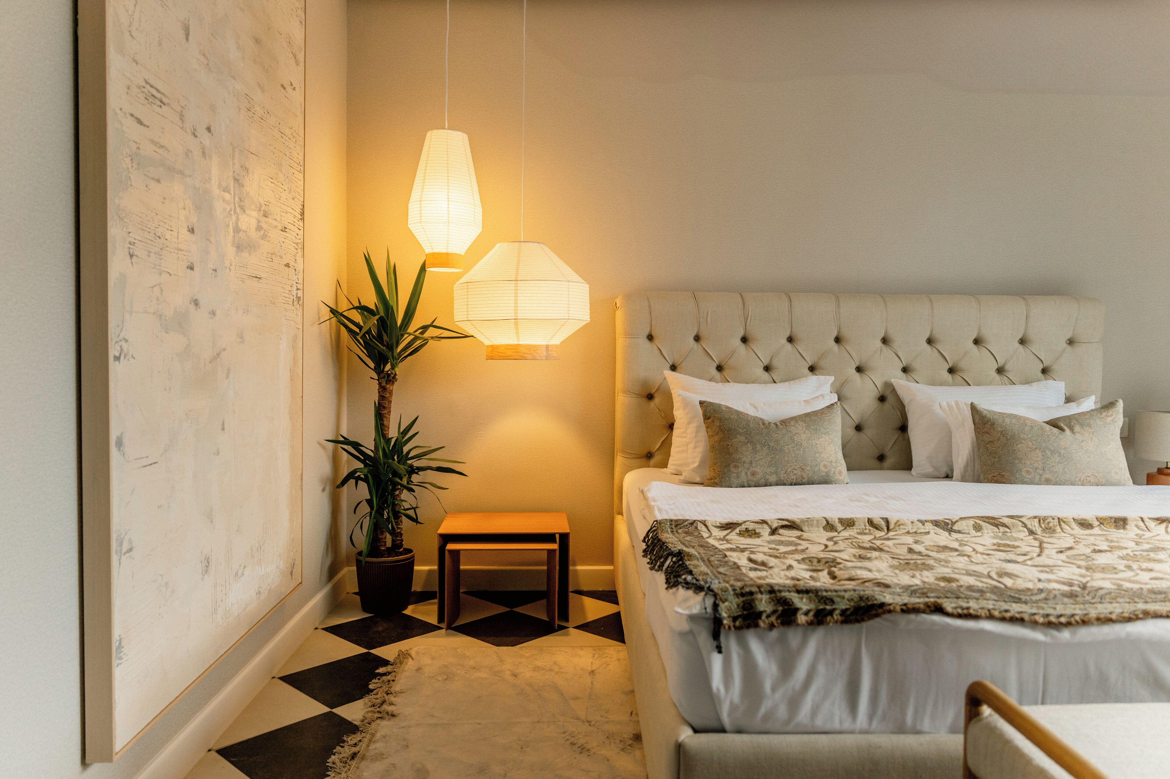 A bedroom with a tufted headboard, a bed with multiple pillows, a small table, and two hanging lamps.