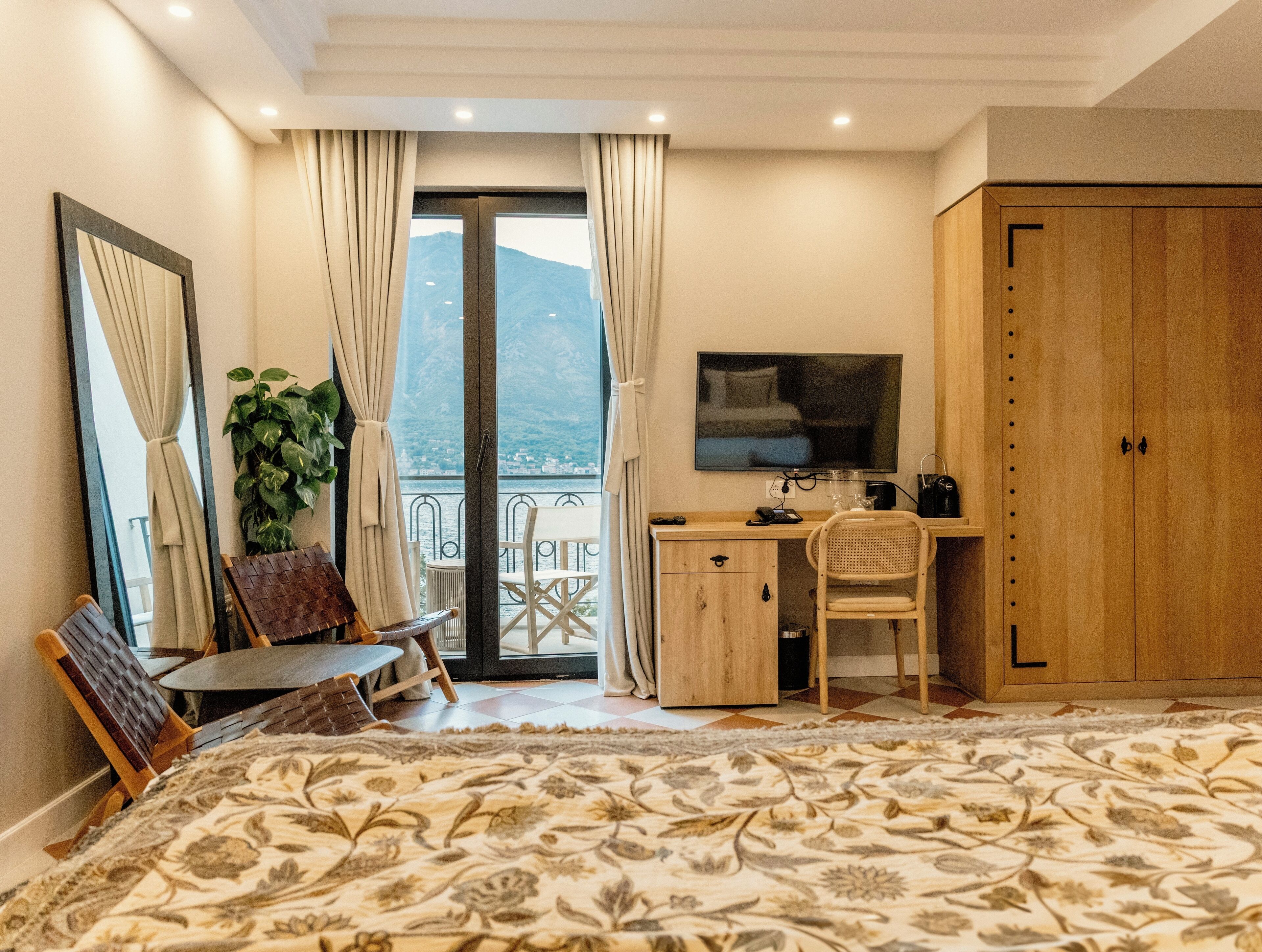 A bedroom with a bed, a desk, a chair, a TV, a wardrobe, and a balcony with a view of the mountains.