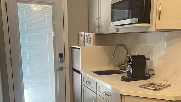 A compact kitchen with white cabinets, a microwave, a coffee maker, and a window with blinds. - Stylish In-law Suite in Frederick (Frederick)