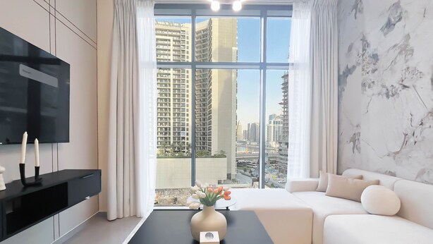 A modern living room with a large window, a flat-screen TV, a black coffee table with a vase of flowers, and a white sofa. - Luxury 2BR in Merano Tower Stunning City Views (Dubai)