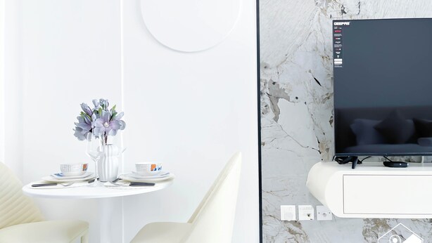 A modern living room with a white TV mounted on a marble wall, a dining table with a vase of flowers, and a sofa. - JVC Hideaway: Studio With Balcony and Amenities (Dubai)