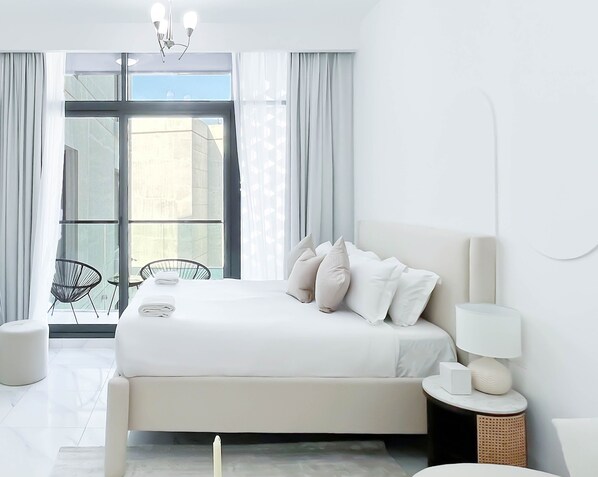 Quarto moderno com uma cama grande, uma mesa de cabeceira com abajur, uma cadeira e vista da cidade através de amplas janelas.