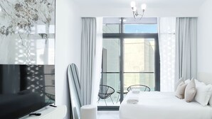 A modern hotel room with a large flat-screen TV, a bed, a seating area with chairs, and a large window with curtains. - JVC Hideaway: Studio With Balcony and Amenities (Dubai)