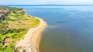 An aerial view of a coastal area with a beach, greenery, and a distant mountain range.