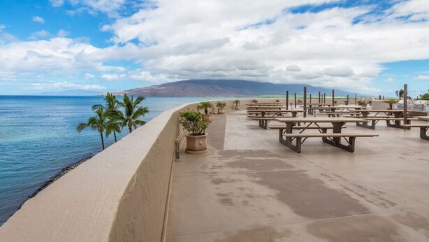 A beachside patio with picnic tables and potted plants, overlooking the ocean and a mountainous landscape.