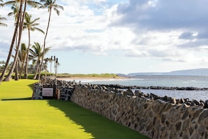 A golf course with a stone wall, palm trees, and a beach in the background. - Hotel-zoned Maui True Paradise - Menehune Shores 227 (Kihei)