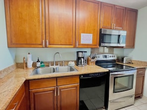 A kitchen with wooden cabinets, a stainless steel sink, a coffee maker, a toaster, and an oven. - Hotel-zoned Maui True Paradise - Menehune Shores 227 (Kihei)