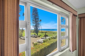 Una habitación con amplios ventanales que ofrecen vista a una playa, coches estacionados y un árbol alto.