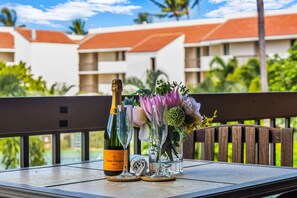 Ein Tisch mit einer Flasche Veuve Clicquot Brut Champagner, zwei Weingläsern und einer Blumenkomposition auf einem Balkon mit Blick auf ein Resort mit Palmen und Gebäuden.