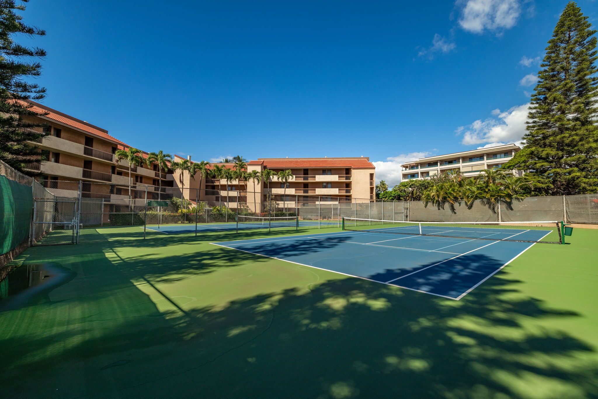 A tennis court with a net, surrounded by a fence, and adjacent to a building with multiple balconies.
