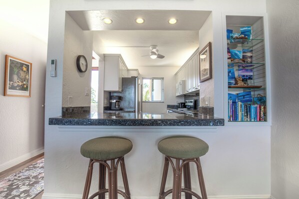 Cocina con barra, dos taburetes, un reloj y una estantería con libros y objetos decorativos.