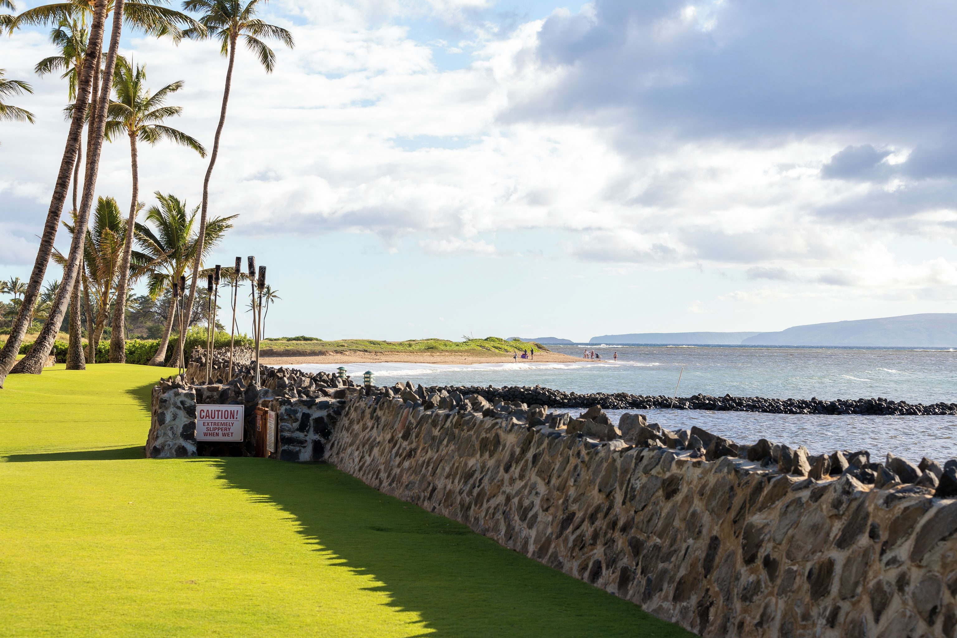 A golf course with a stone wall, palm trees, and a beach in the background.