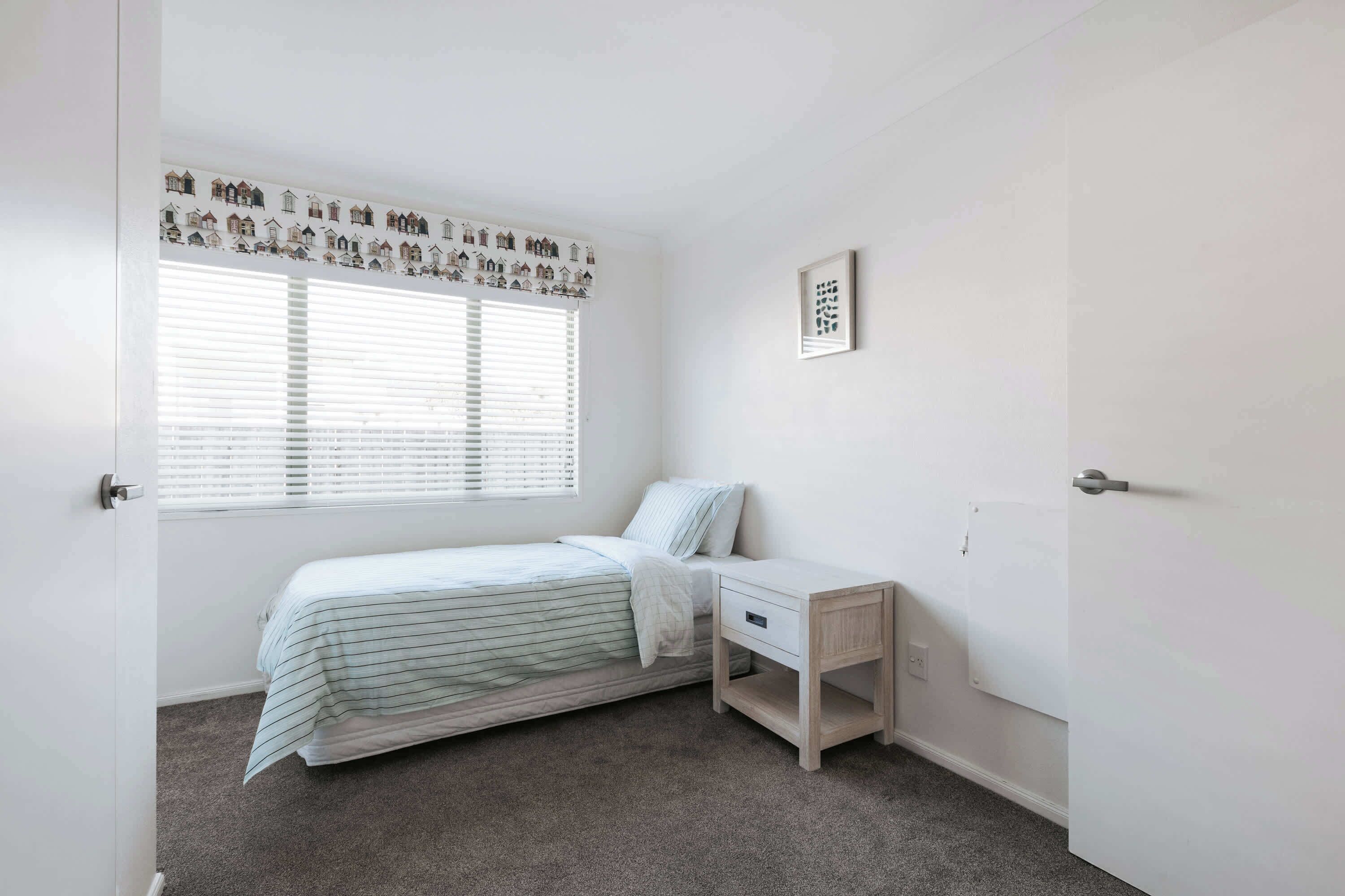 A bedroom with a single bed, a bedside table, a window with blinds, a wall-mounted picture, and a door.