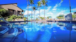 Una piscina azul cristalina con un borde de baldosas, rodeada de palmeras y sillas de descanso, con un edificio al fondo.