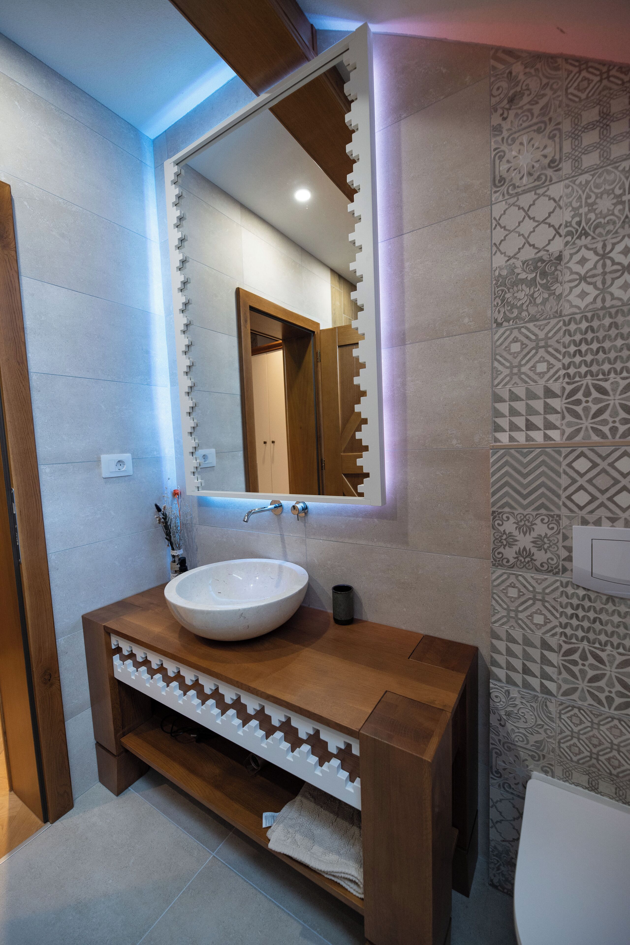 A bathroom with a wooden vanity, a white sink, and a mirror with a geometric frame.