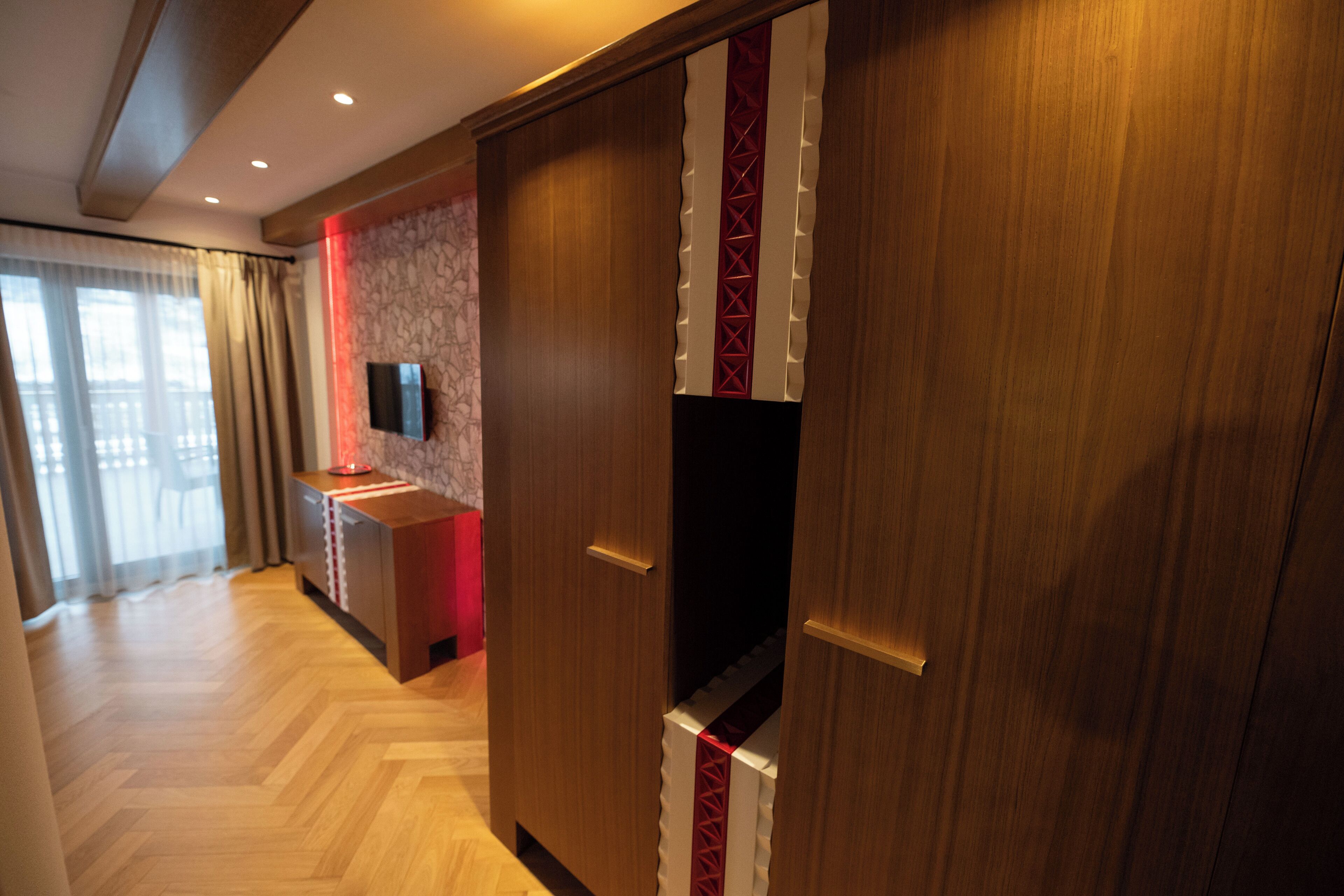A hotel room with a wooden floor, a red accent wall, a flat-screen TV mounted on the wall, and a wardrobe with a red and white design.