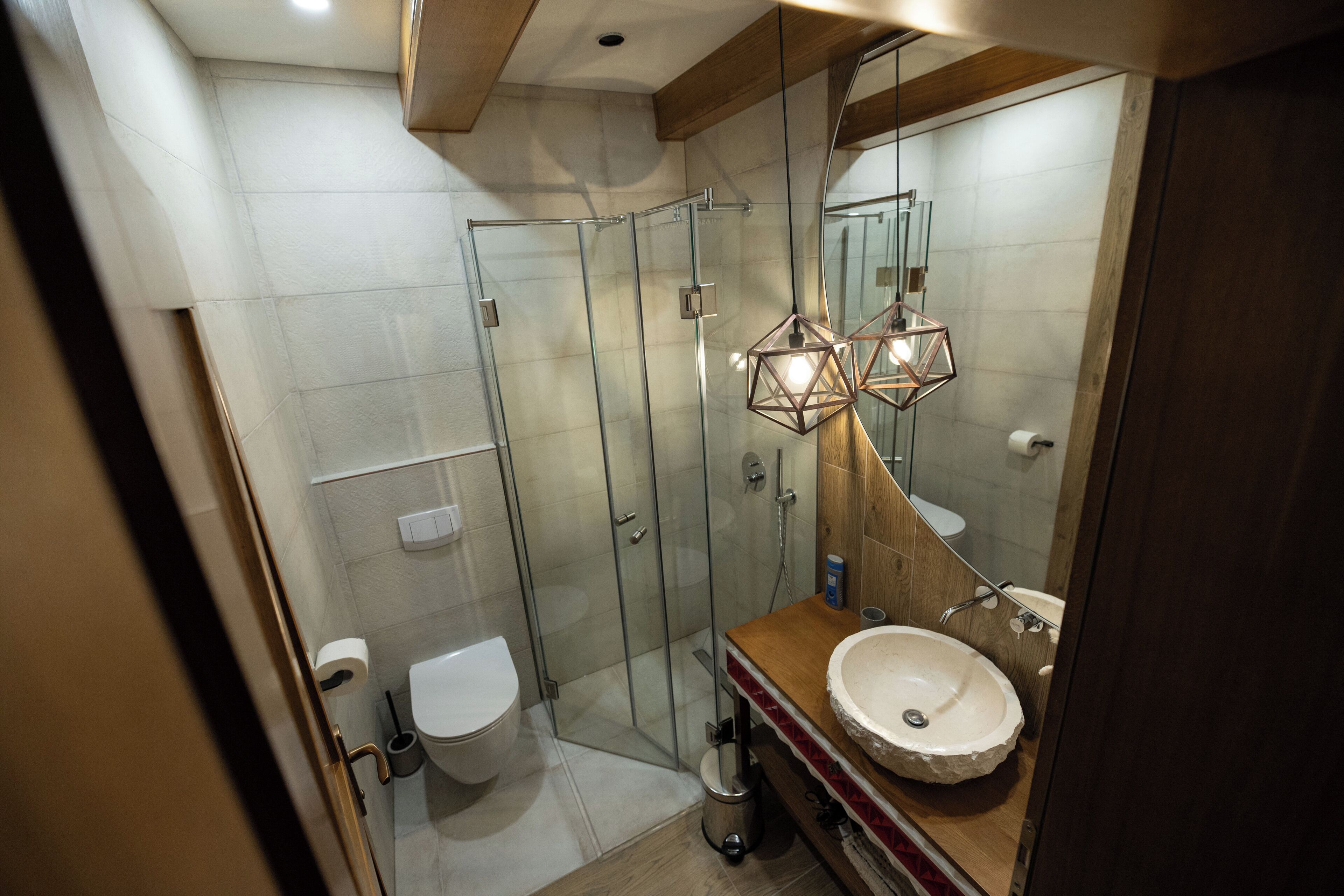 A modern bathroom with a glass shower enclosure, a wooden vanity with a round sink, and a geometric pendant light.