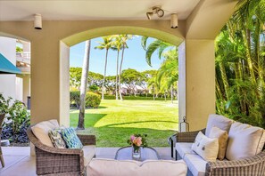 A covered outdoor seating area with wicker furniture, a view of a well-manicured lawn, and palm trees.