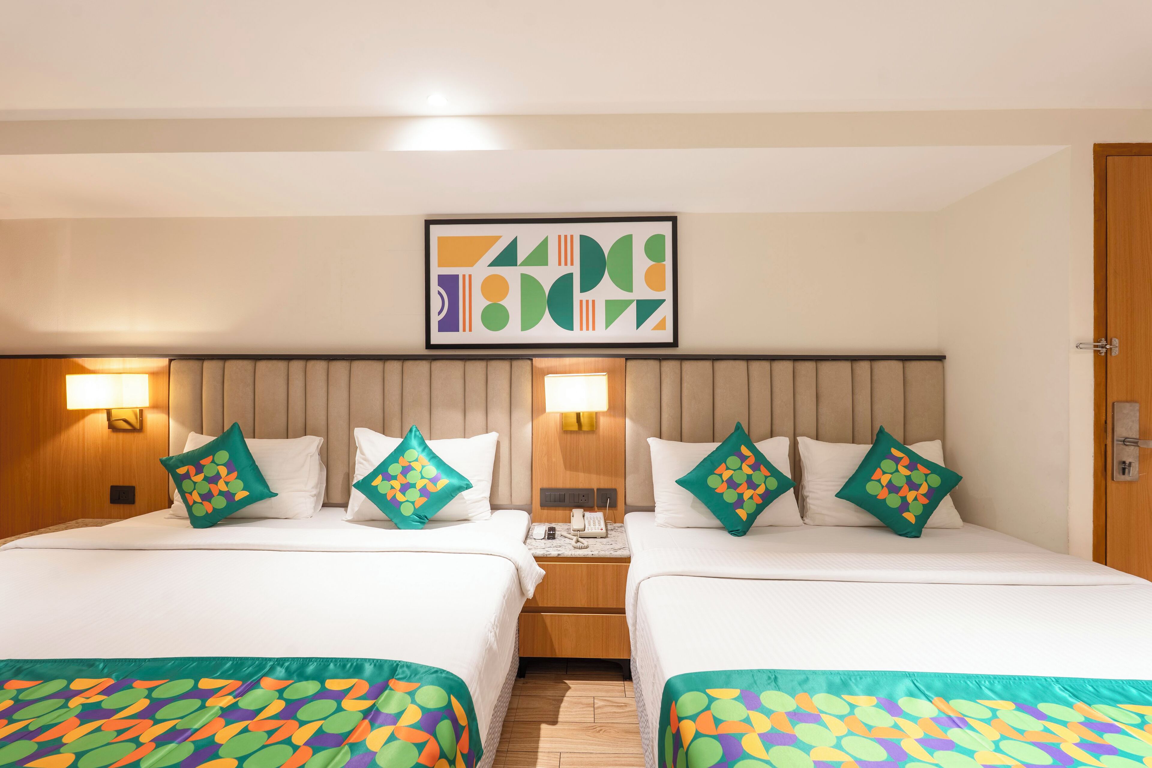 A hotel room with two beds, each with a patterned bedspread, a headboard, bedside lamps, and a framed abstract artwork above the beds.