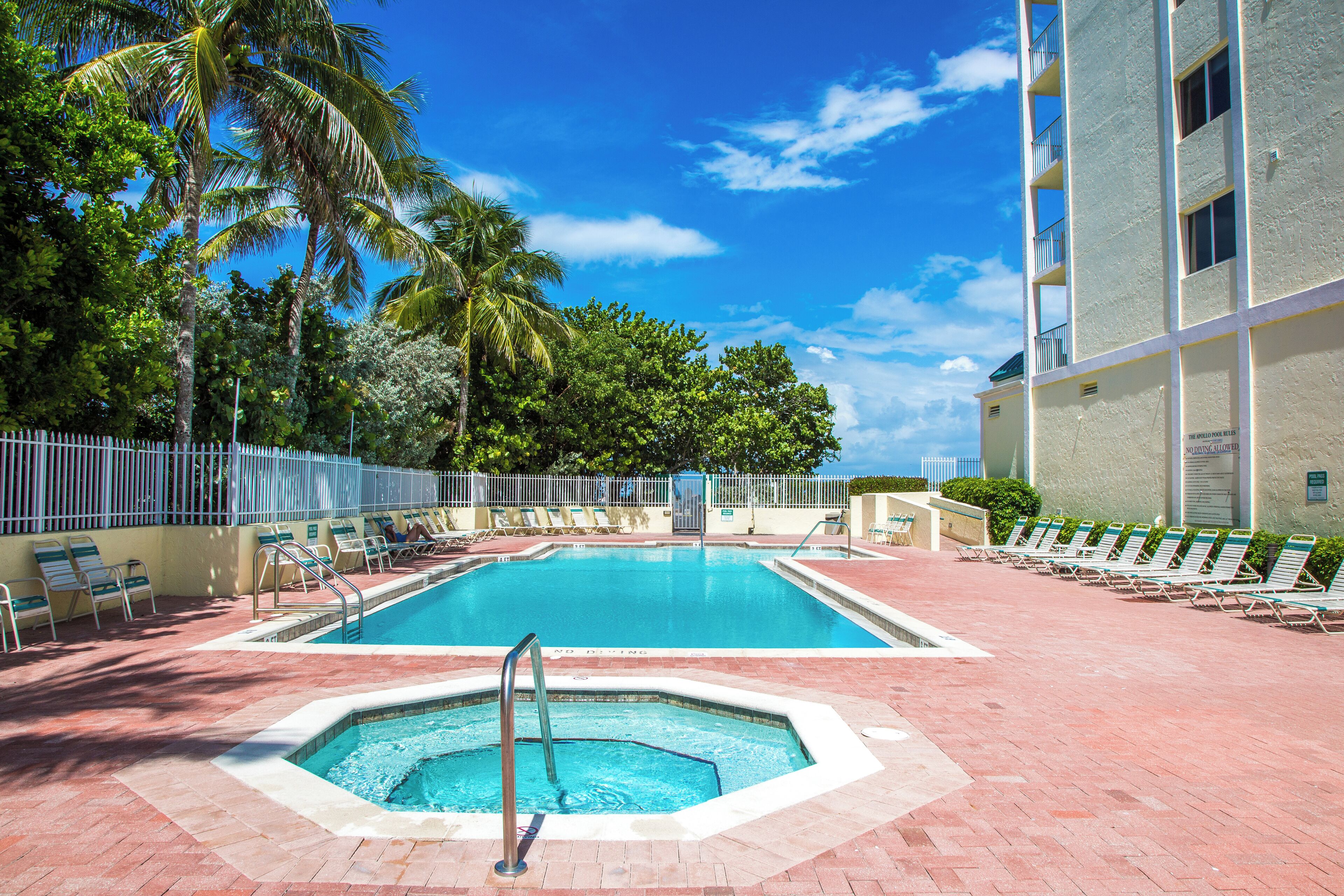 A swimming pool with lounge chairs and a hot tub, surrounded by palm trees and a building.